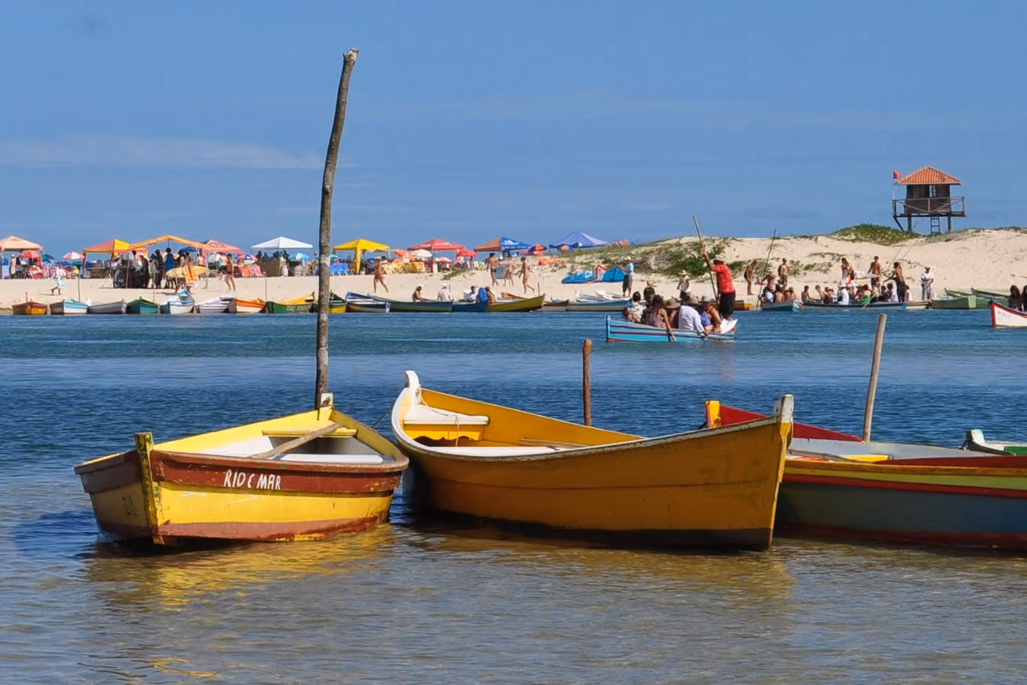 Botes de colores en el Rio da Madre, en Guarda do Embaú, con un bote que lleva gente hasta la playa y el puesto de guardavidas en los médanos al fondo