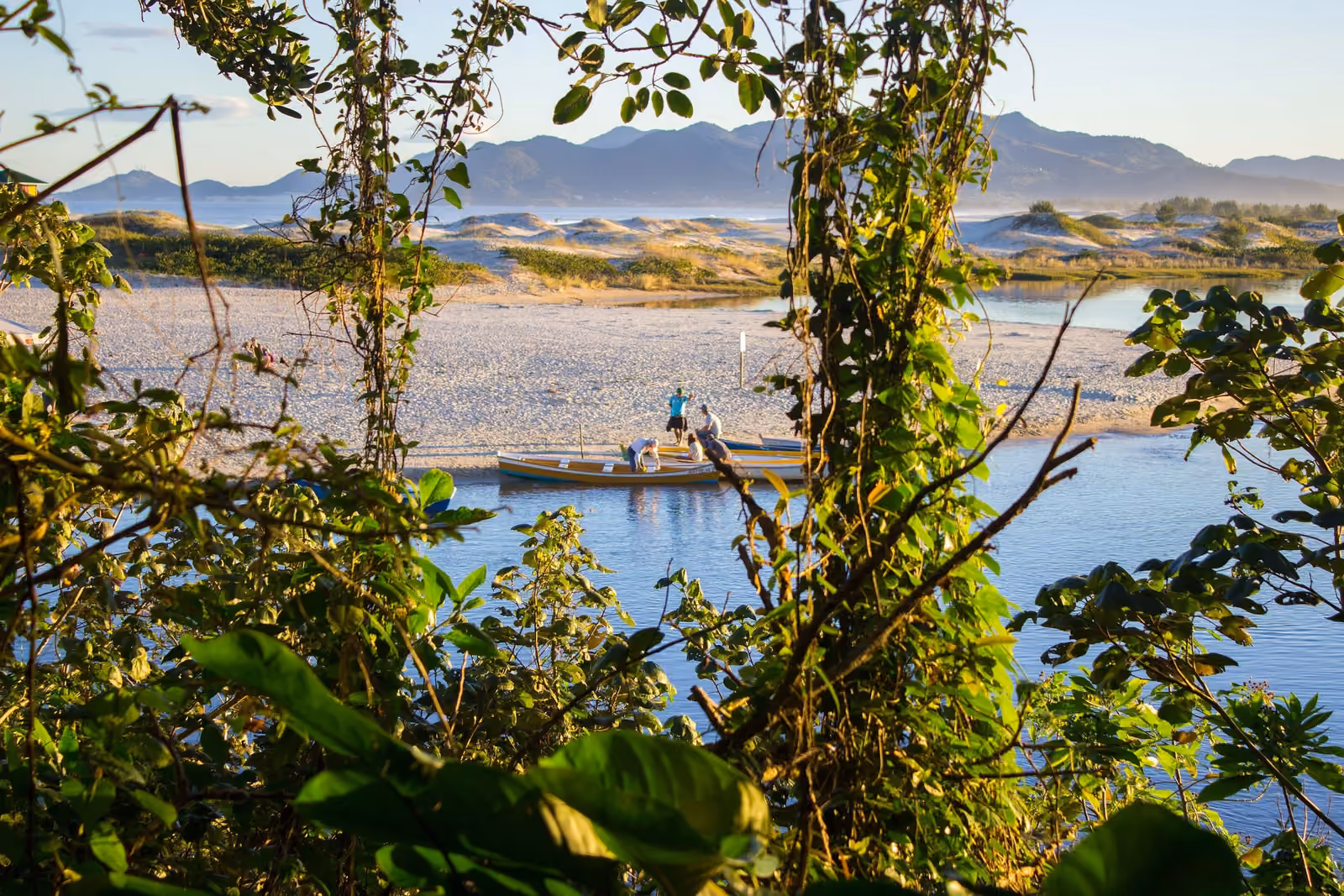 Botes en la orilla del Rio da Madre vistos entre la vegetación, con la arena, los médanos y la sierra al fondo, al atardecer