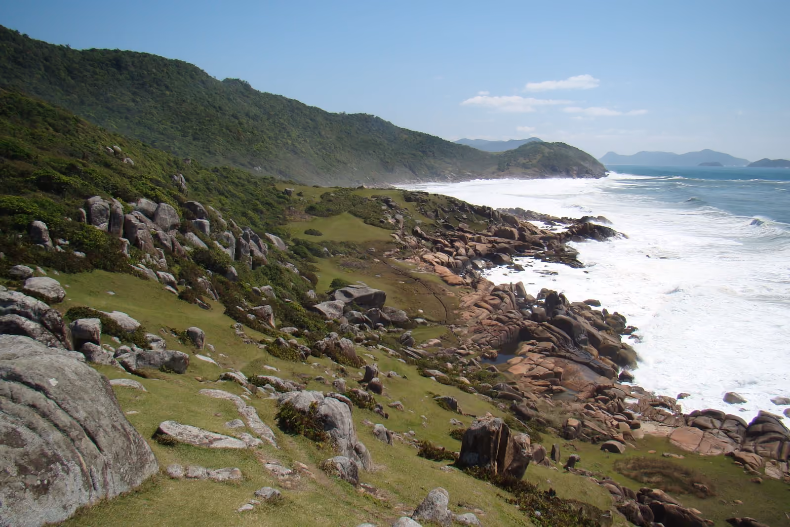Costa rocosa de Guarda do Embaú, con piedras entre el pasto, el morro cubierto de vegetación y el mar rompiendo blanco contra las rocas