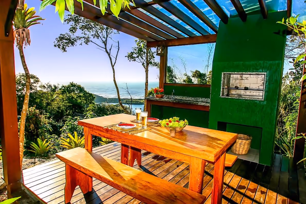 Deck com mesa de madeira e churrasqueira da Morada da Lagoa, na Praia do Rosa, com vista para o mar entre a mata