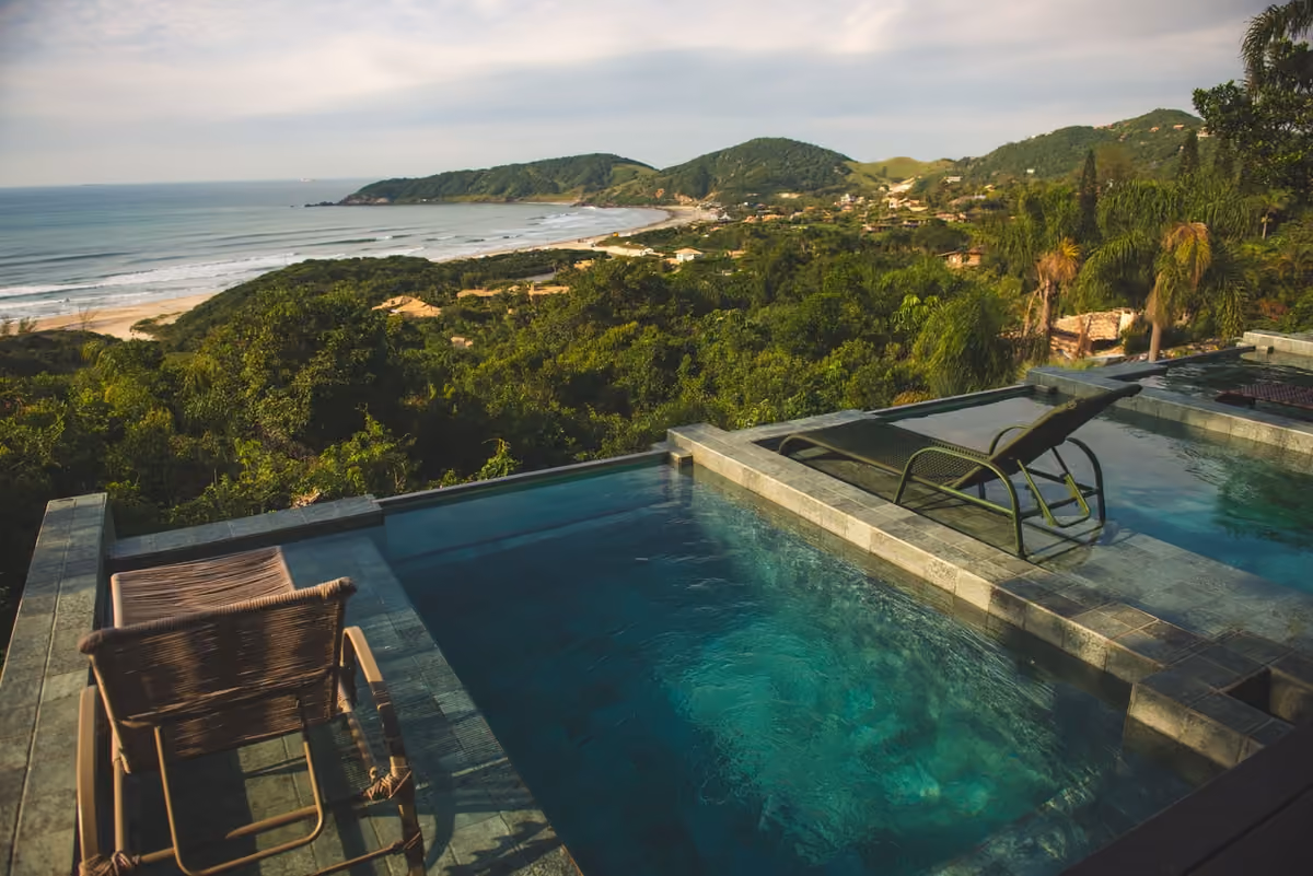 Piscina de borda infinita da Pousada Rosa Norte, na Praia do Rosa, com espreguiçadeiras e vista para a enseada e os morros cobertos de mata