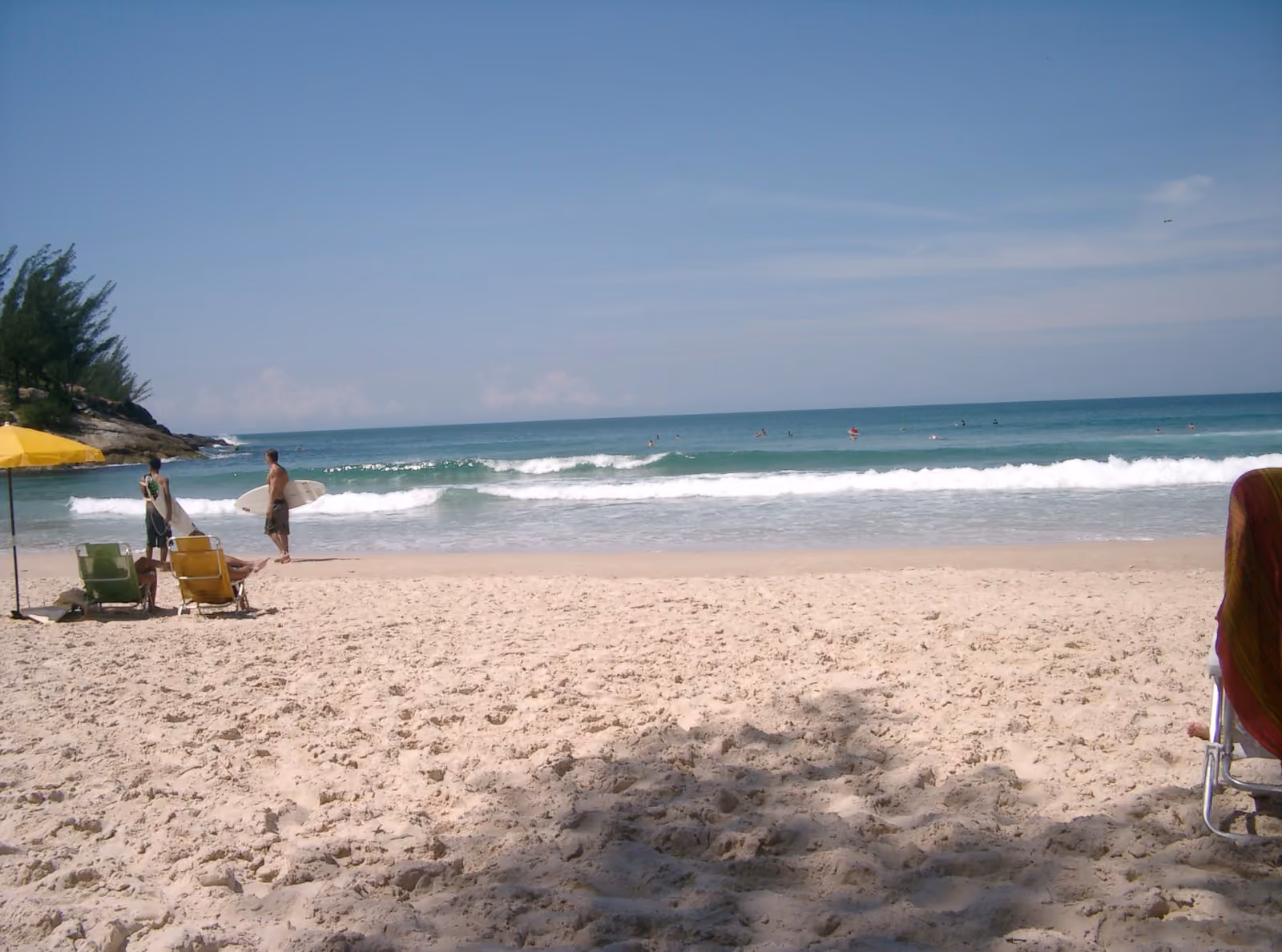 Surfistas con tabla en la arena de la Praia da Ferrugem, junto a una sombrilla y reposeras, con olas rompiendo