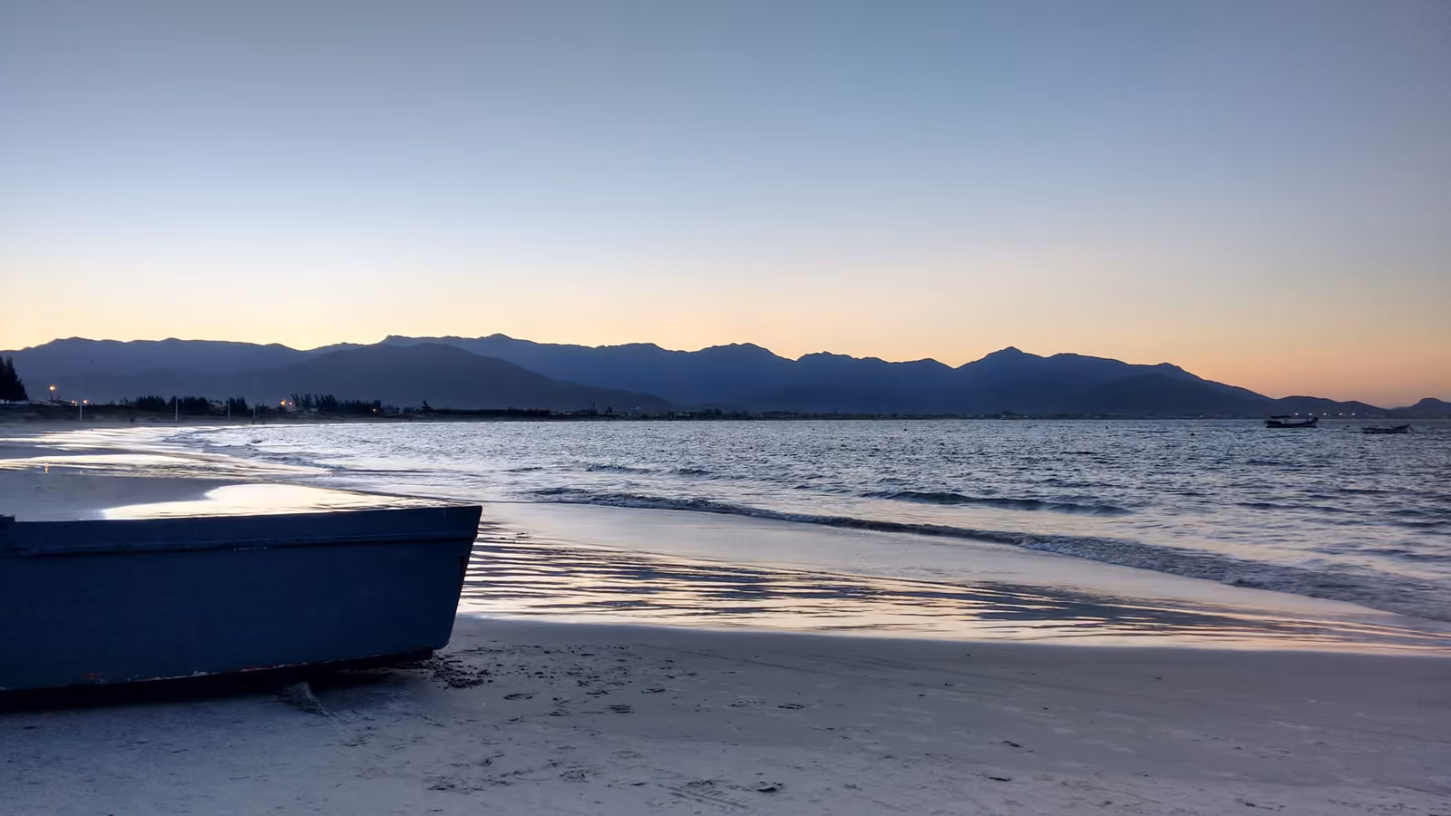 Praia da Pinheira ao entardecer, com um barco na areia, a orla iluminada e a serra ao fundo