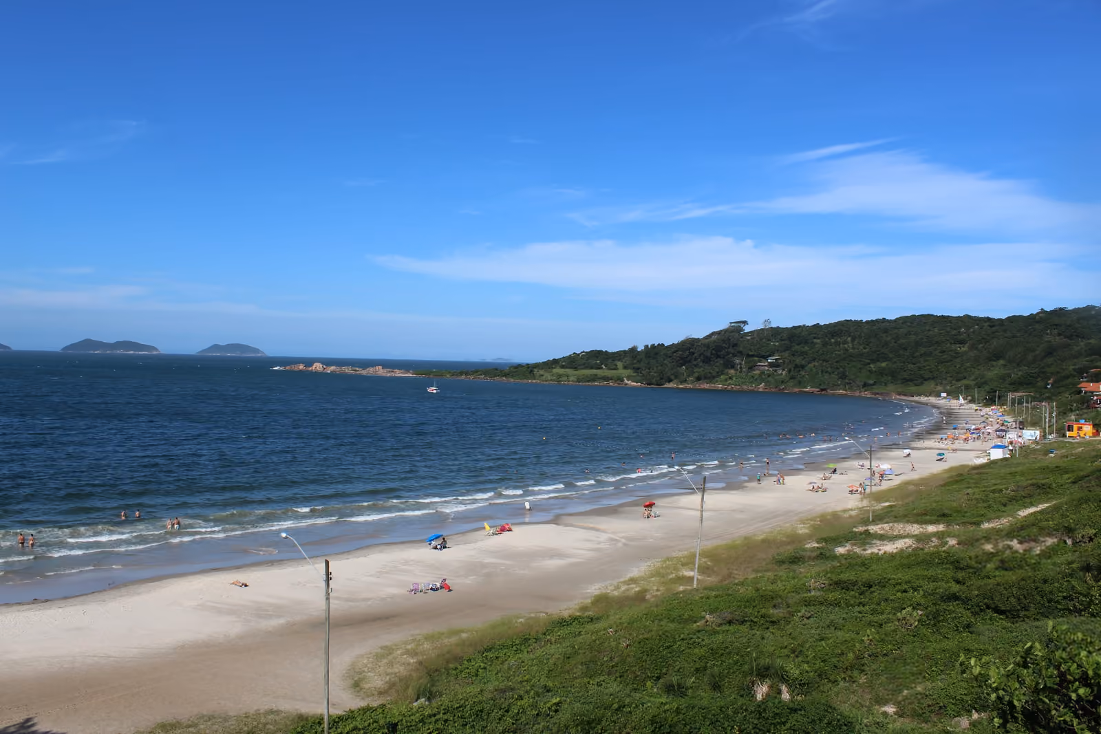 Praia de Cima, na Pinheira, vista do alto da vegetação, com banhistas e guarda-sóis na areia, o morro com mata à direita e ilhas no horizonte