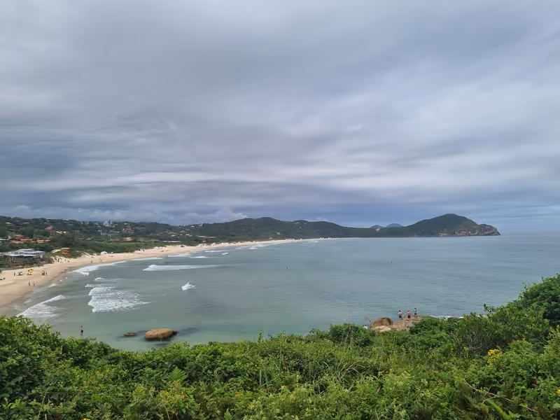 Enseada da Praia do Rosa vista do alto do costão, com a faixa de areia, o mar e o morro ao fundo, num dia nublado